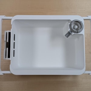 May include: A white plastic ice maker component with a rectangular ice tray and a gray dispenser. The tray is set within a white frame. The dispenser is on the upper right corner of the tray.