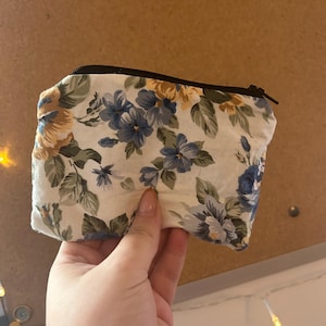 Puede incluir: Pequeño neceser rectangular con estampado floral y cremallera negra. La tela presenta un patrón de flores azules, amarillas y verdes sobre un fondo blanco. El neceser se sostiene sobre un fondo de tono neutro.