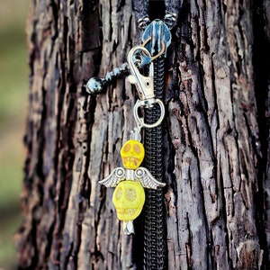 Könnte beinhalten: Ein silberner Schlüsselanhänger mit einem gelben Totenkopf-Anhänger. Der Anhänger besteht aus zwei Totenköpfen mit Flügeln. Der Schlüsselanhänger hängt an einem Baumstamm.