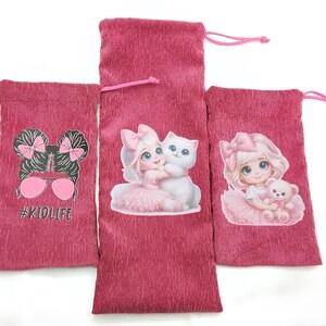 Könnte beinhalten: Drei rosa Kordelzugbeutel mit verschiedenen Designs. Eine Tasche zeigt eine Grafik eines Mädchens mit Sonnenbrille und dem Text "#KIDLIFE". Eine andere Tasche hat ein Mädchen, das eine weiße Katze umarmt. Die dritte Tasche hat ein Mädchen, das einen Teddybären hält.