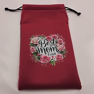 Puede incluir: Una bolsa roja con cordón con el texto "Best Mom Ever" rodeado por una corona floral de rosas rosas y hojas verdes. La bolsa tiene una superficie texturizada y cordones negros.
