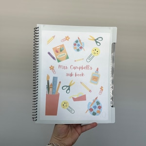 May include: A white binder with a colorful design of school supplies, including crayons, scissors, paintbrushes, and a paint palette. The binder has the text "Mrs. Campbell's Sub-Book" printed on it.