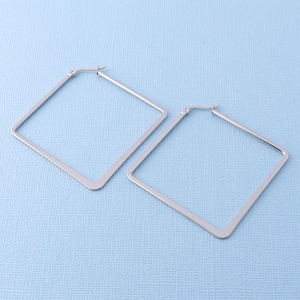 May include: A pair of silver-toned square hoop earrings featuring a thin, flat metal construction with a hinged post closure at the top corner. The earrings have a smooth, polished finish and a geometric shape, presented as two separate pieces.