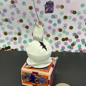 Puede incluir: Una taza de té de cerámica blanca con una bolsita de té dentro, sentada sobre una pequeña caja con un diseño colorido. Una cuchara de plata está al lado de la taza. Un pequeño amuleto con las palabras "Boss B*tch Tea Cup" cuelga de un lazo de alambre.