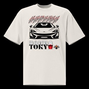 Könnte beinhalten: Cremefarbenes T-Shirt mit einer schwarz-weißen Illustration eines Sportwagens. Über dem Auto befindet sich ein abstraktes rotes und schwarzes Design. Unter dem Auto steht das Wort "TOKYO" in Schwarz, mit einer rot-weißen Grafik und einer Zielflagge.