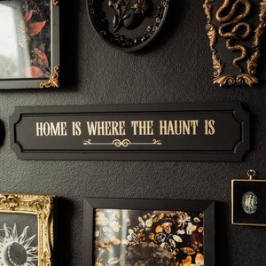A black and gold wall sign that reads 'home is where the haunt is' hung on the wall as part of a gothic maximalism gallery