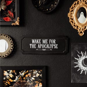 Black, rectangular, Victorian‑style wall plaque with text saying ‘Wake me for the apocalypse’ in white Gothic lettering; hangs by a sawtooth hook. Displayed as part of a gothic, maximalist gallery wall.