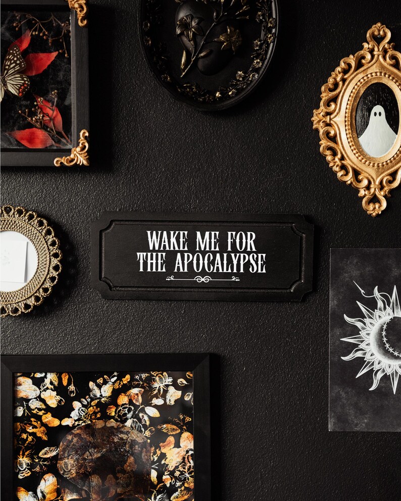 Black, rectangular, Victorian‑style wall plaque with text saying ‘Wake me for the apocalypse’ in white Gothic lettering; hangs by a sawtooth hook. Displayed as part of a gothic, maximalist gallery wall.