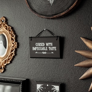 May include: Black and white sign with the text "Cursed with impeccable taste" hanging on a black wall.