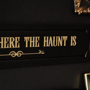 May include: Black sign with gold lettering that reads "Where the haunt is".