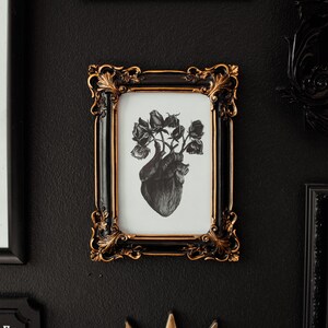 May include: A black and white print of a heart with roses growing from it, framed in a black and gold ornate frame. The frame is hanging on a black wall.