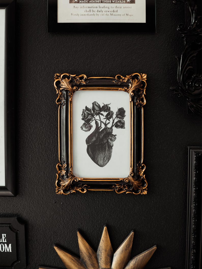 May include: A black and white print of a heart with roses growing from it, framed in a black and gold ornate frame. The frame is hanging on a black wall.