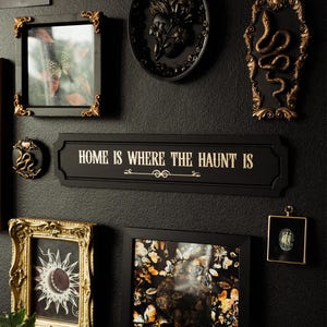 May include: A black wall gallery featuring a variety of framed artwork, including a skull with butterflies, a snake, and a floral design. A black sign with gold lettering reads "Home is where the haunt is".
