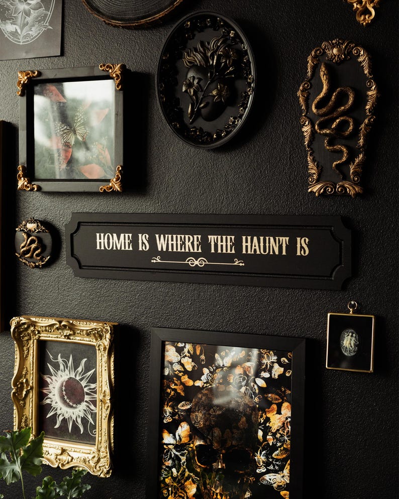 May include: A black wall gallery featuring a variety of framed artwork, including a skull with butterflies, a snake, and a floral design. A black sign with gold lettering reads "Home is where the haunt is".