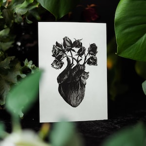 A black and white print of a hand-drawn anatomical heart blooming with wilted roses as though it were a vase. Surrounded by dark green leaves and shadows. Ideal for gothic or anatomy-themed decor.