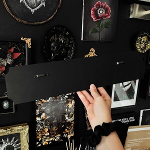 May include: A black metal sign with two small black clips on the back. The sign is hanging on a black wall with various framed artwork and decor.