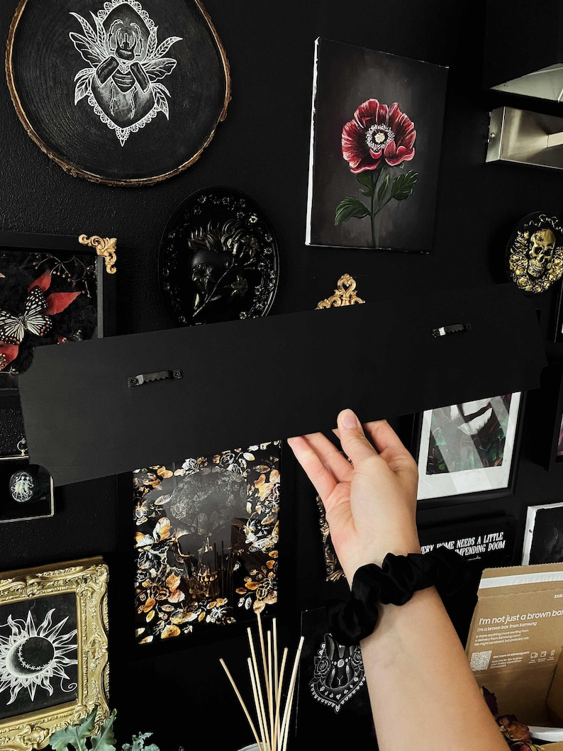 May include: A black metal sign with two small black clips on the back. The sign is hanging on a black wall with various framed artwork and decor.