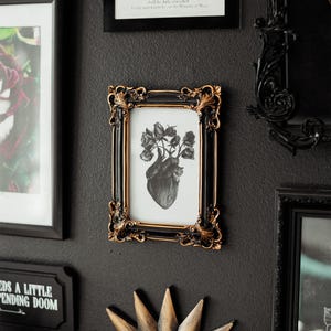 May include: A black framed print with a gold trim, featuring a black and white anatomical heart with roses growing from it. The print is hanging on a black wall with other framed prints and a gold mirror.
