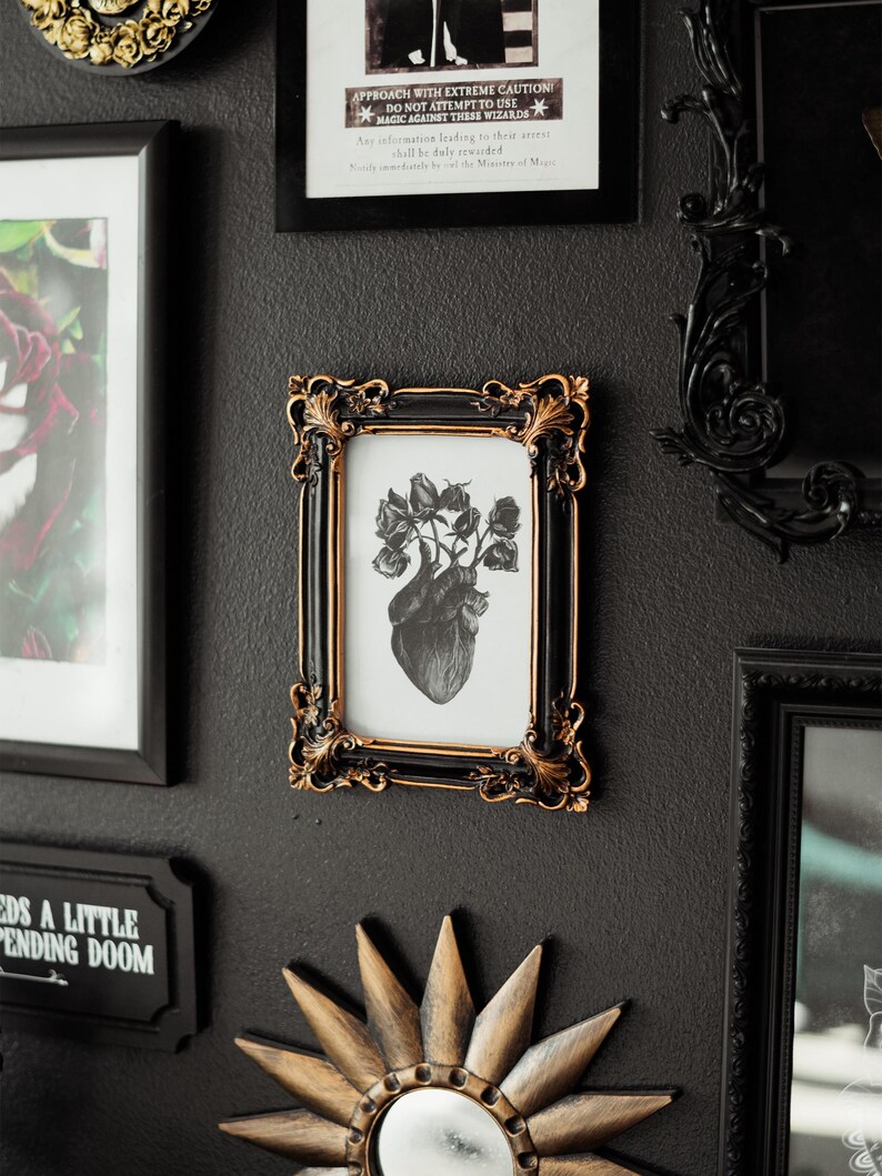 May include: A black framed print with a gold trim, featuring a black and white anatomical heart with roses growing from it. The print is hanging on a black wall with other framed prints and a gold mirror.
