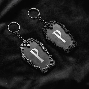 May include: Two silver keychains with ornate frames and black backgrounds. Each frame features a white letter "P" in the centre.