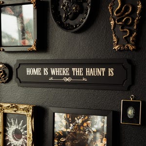 May include: A black and gold wall decor sign with the text "Home is where the haunt is". The sign is surrounded by other decorative items, including a black framed picture with a skull and flowers, a black framed picture with a snake, and a black framed picture with a heart.