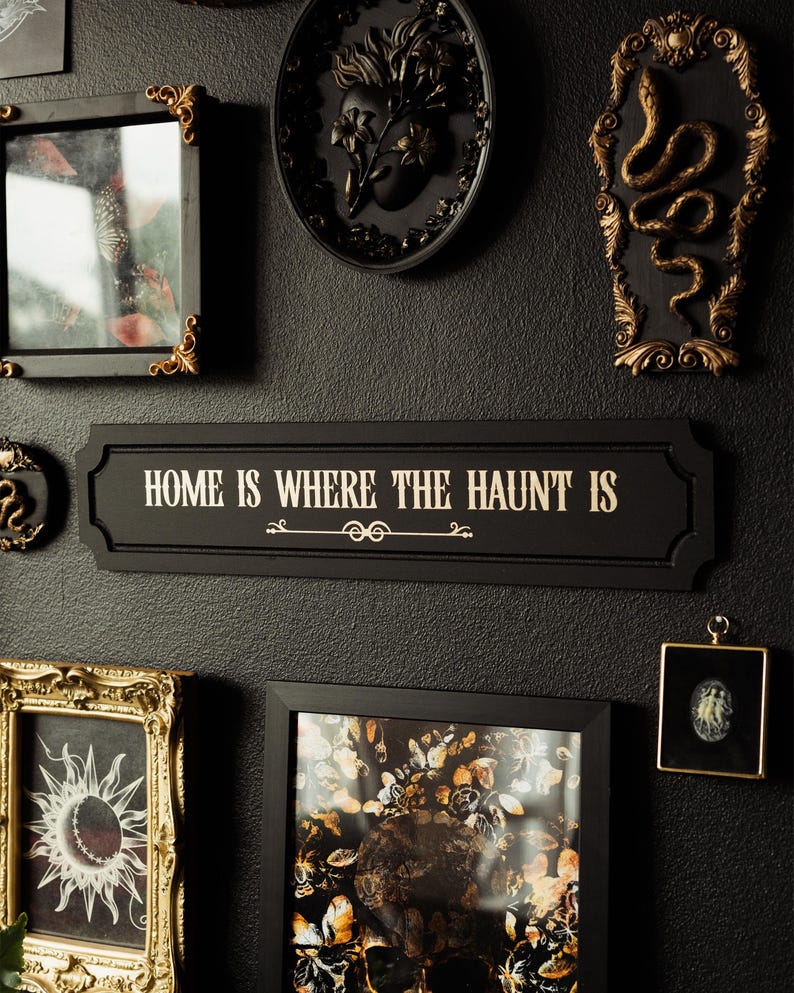 May include: A black and gold wall decor sign with the text "Home is where the haunt is". The sign is surrounded by other decorative items, including a black framed picture with a skull and flowers, a black framed picture with a snake, and a black framed picture with a heart.