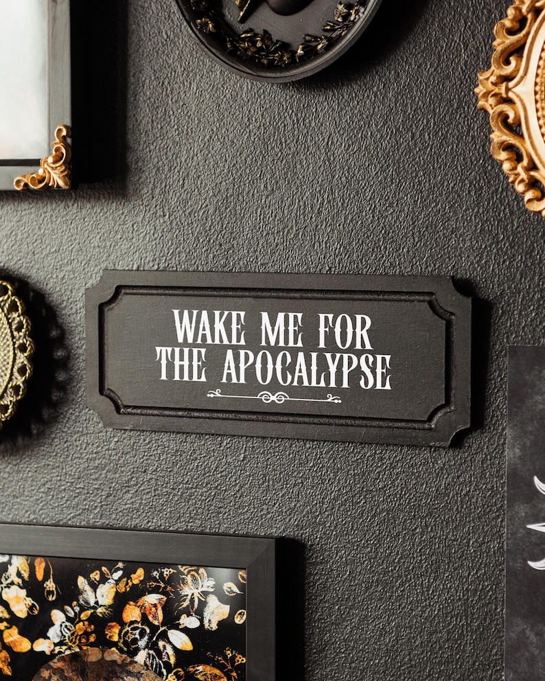 Black, rectangular, Victorian‑style wall plaque with text saying ‘Wake me for the apocalypse’ in white Gothic lettering; hangs by a sawtooth hook. Displayed as part of a gothic, maximalist gallery wall.