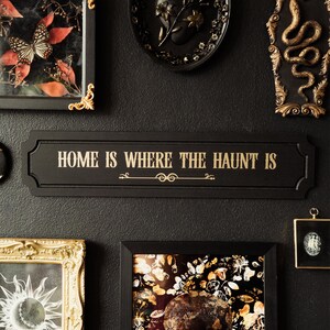 May include: Black and gold framed wall decor with a black sign that reads "Home is where the haunt is". The sign is surrounded by other framed artwork, including a butterfly, a skull, and a snake.