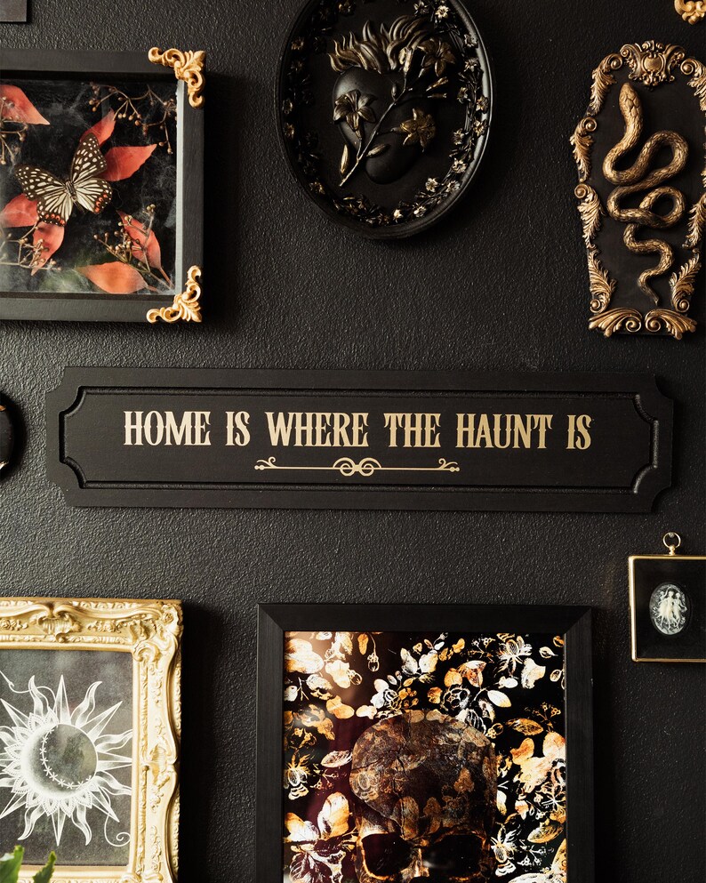 May include: Black and gold framed wall decor with a black sign that reads "Home is where the haunt is". The sign is surrounded by other framed artwork, including a butterfly, a skull, and a snake.