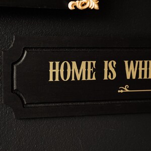 May include: Black wooden sign with gold lettering that reads "HOME IS WHERE THE HEART IS".