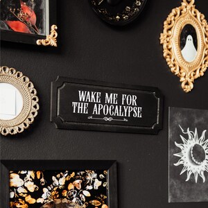Black, rectangular, Victorian‑style wall plaque with text saying ‘Wake me for the apocalypse’ in white Gothic lettering; hangs by a sawtooth hook. Displayed as part of a gothic, maximalist gallery wall.