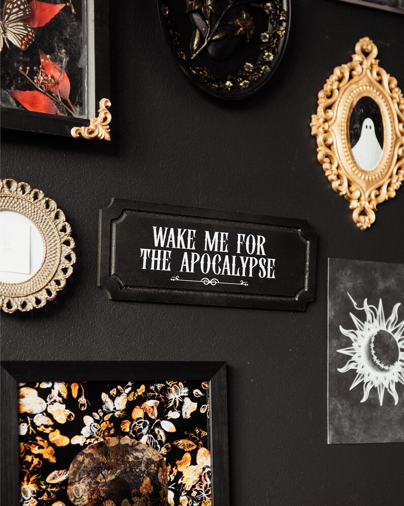 Black, rectangular, Victorian‑style wall plaque with text saying ‘Wake me for the apocalypse’ in white Gothic lettering; hangs by a sawtooth hook. Displayed as part of a gothic, maximalist gallery wall.