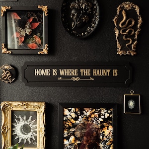 May include: A black and gold framed sign with the text "Home is where the haunt is" in gold lettering. The sign is surrounded by other framed artwork, including a black and gold framed skull with butterflies, a black and gold framed butterfly, a black and gold framed snake, and a black and gold framed sun.