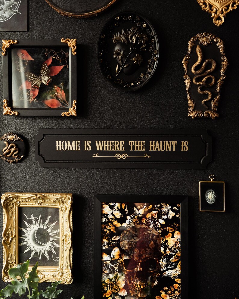 May include: A black and gold framed sign with the text "Home is where the haunt is" in gold lettering. The sign is surrounded by other framed artwork, including a black and gold framed skull with butterflies, a black and gold framed butterfly, a black and gold framed snake, and a black and gold framed sun.
