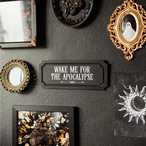 Black, rectangular, Victorian‑style wall plaque with text saying ‘Wake me for the apocalypse’ in white Gothic lettering; hangs by a sawtooth hook. Displayed as part of a gothic, maximalist gallery wall.