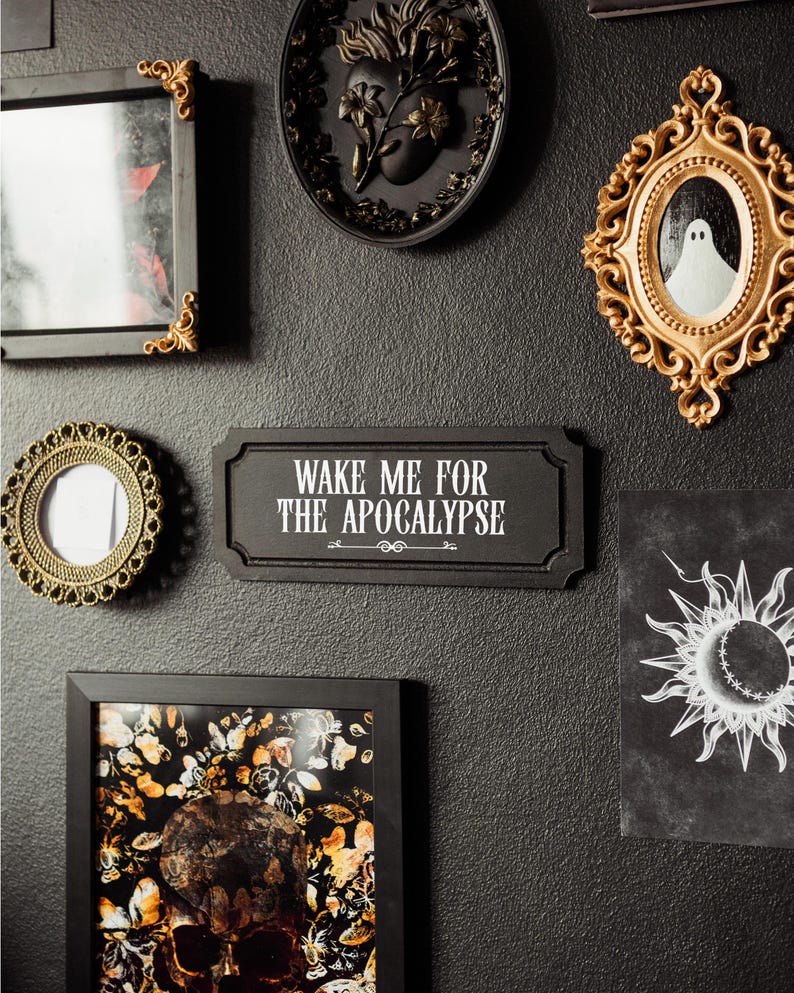Black, rectangular, Victorian‑style wall plaque with text saying ‘Wake me for the apocalypse’ in white Gothic lettering; hangs by a sawtooth hook. Displayed as part of a gothic, maximalist gallery wall.
