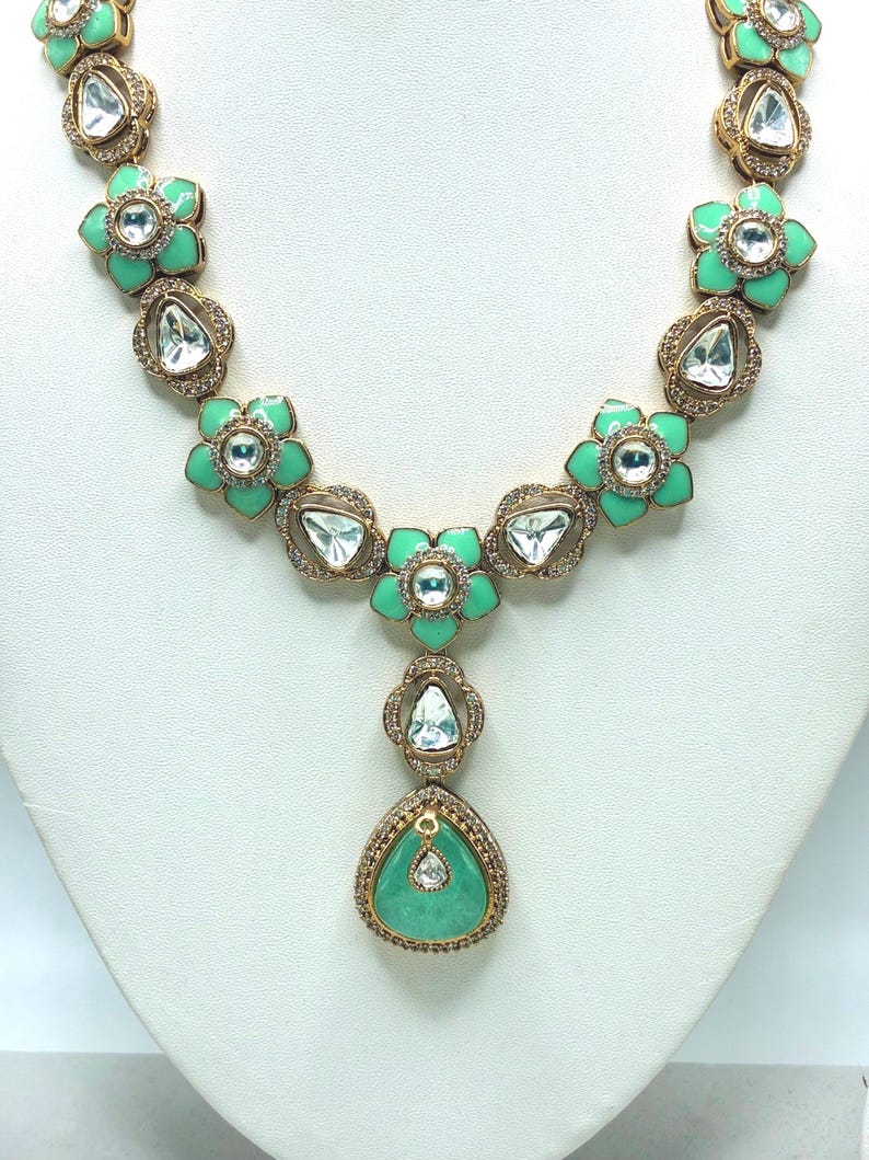 May include: A gold-toned necklace featuring a floral design with mint green enamel petals and clear, faceted gemstones. The necklace has a central pendant with a teardrop-shaped, mint green stone, surrounded by smaller clear stones.
