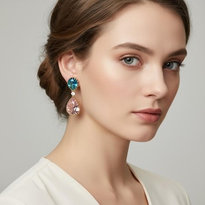 Könnte beinhalten: Ein Paar elegante Ohrhänger mit teal- und rosafarbenen Edelsteinen in einer goldfarbenen Fassung. Die Ohrringe haben eine Tropfenform, mit einem kleinen weißen Edelstein, der die beiden größeren Steine trennt.