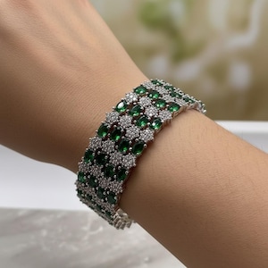 May include: A silver bracelet with three rows of oval-cut emerald green gemstones and small, round, clear gemstones. The bracelet is worn on a wrist.