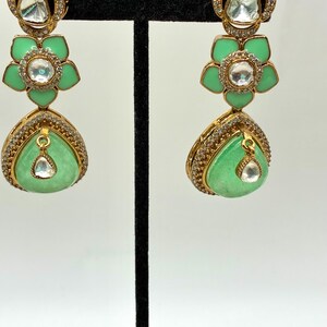 May include: Gold-toned drop earrings with a floral design. The earrings feature a light green, teardrop-shaped stone and a flower-shaped element in the middle. The earrings are adorned with clear, sparkling accents.