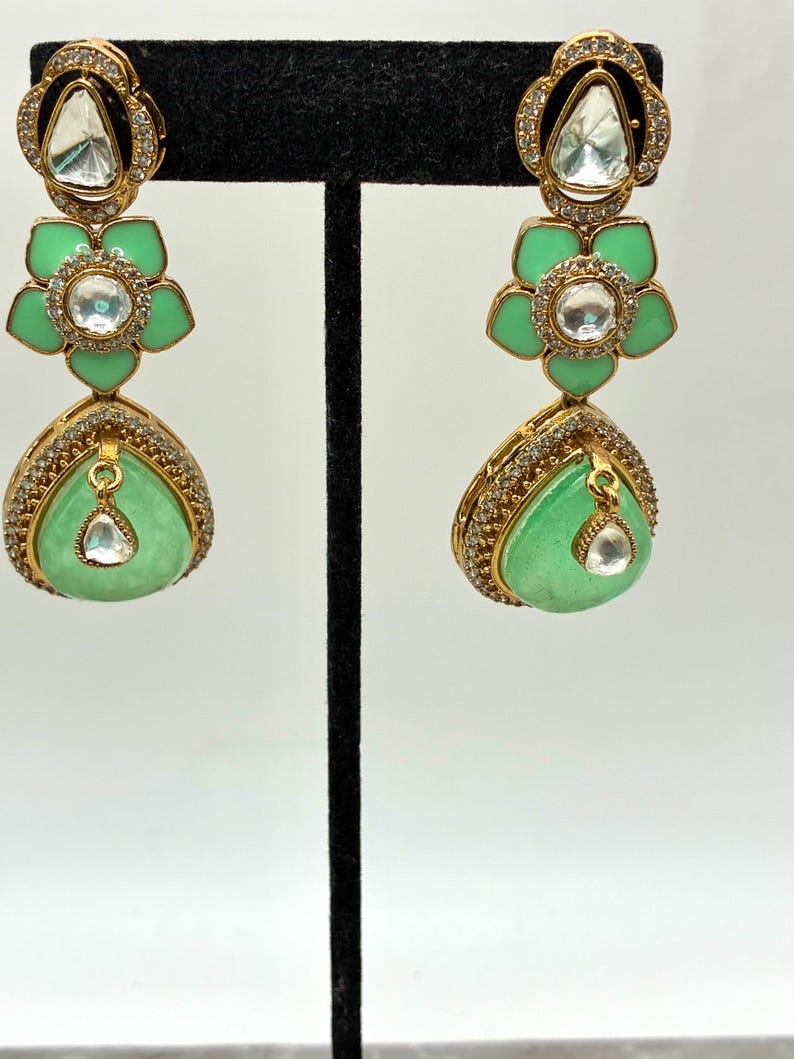 May include: Gold-toned drop earrings with a floral design. The earrings feature a light green, teardrop-shaped stone and a flower-shaped element in the middle. The earrings are adorned with clear, sparkling accents.
