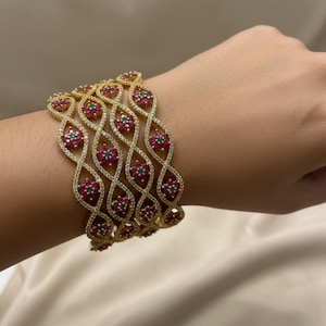 May include: Gold-toned bangles with a repeating floral design. Each bangle features a series of red and green gemstones set within a swirling, openwork pattern, accented with clear crystals. The bracelets are stacked on top of each other.