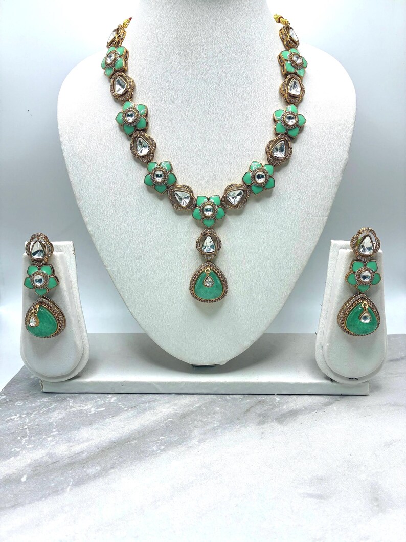 May include: A mint green and gold-toned necklace and earring set. The necklace features flower-shaped pendants with clear stone accents. The earrings mirror the necklace design, with a teardrop-shaped green stone at the bottom. The jewelry is displayed on a white bust.