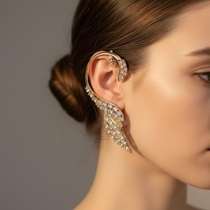 May include: A gold-toned ear cuff featuring a curved, leaf-inspired design that wraps around the outer edge of the ear. The piece is encrusted with numerous small, clear, sparkling crystals and includes a small, cutout leaf detail at the top.