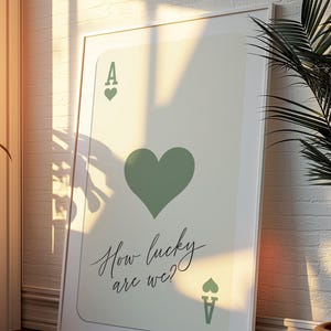 May include: A white framed print of an ace of hearts playing card with a green heart and the text "How lucky are we?"