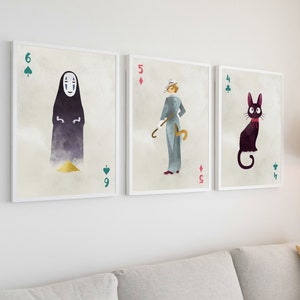 May include: Three framed art prints featuring watercolor illustrations of characters from animated films. The prints resemble playing cards, with a No-Face character, a cat-like figure, and a character with a cane. The frames are white, and the background is a neutral color.