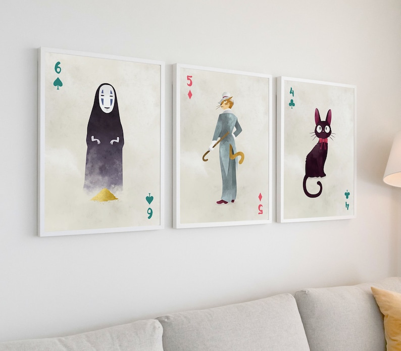 May include: Three framed art prints featuring watercolor illustrations of characters from animated films. The prints resemble playing cards, with a No-Face character, a cat-like figure, and a character with a cane. The frames are white, and the background is a neutral color.