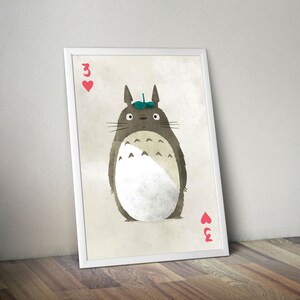 May include: A white framed print of a cartoon Totoro character from the movie My Neighbor Totoro. The character is wearing a green leaf on its head and is holding a playing card with the number 3 of hearts. The background is a light beige color.