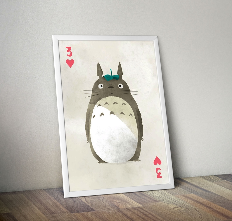 May include: A white framed print of a cartoon Totoro character from the movie My Neighbor Totoro. The character is wearing a green leaf on its head and is holding a playing card with the number 3 of hearts. The background is a light beige color.