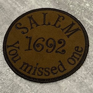 May include: A brown round patch with black embroidered text that reads "Salem 1692 You missed one".